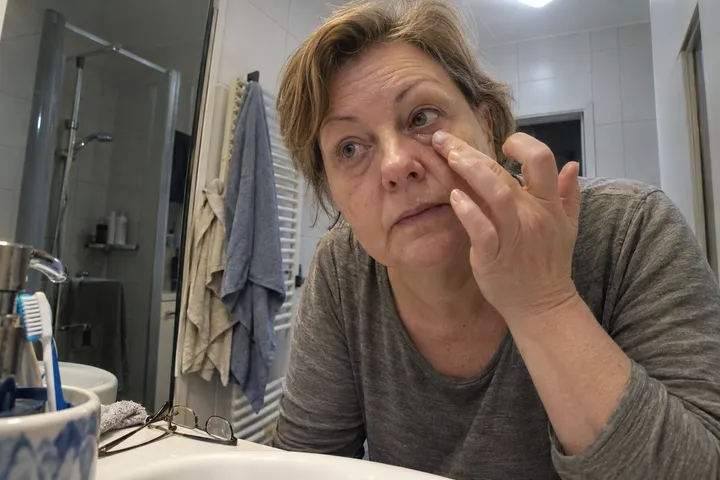 Frau im grauen Langarmshirt tupft sich morgens im Bad unter das Auge, die Augen wirken müde